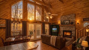 L’intérieur d’un chalet accueillant, avec un coin repas, une cheminée, un mur en pierre et une grande fenêtre donnant sur les arbres.