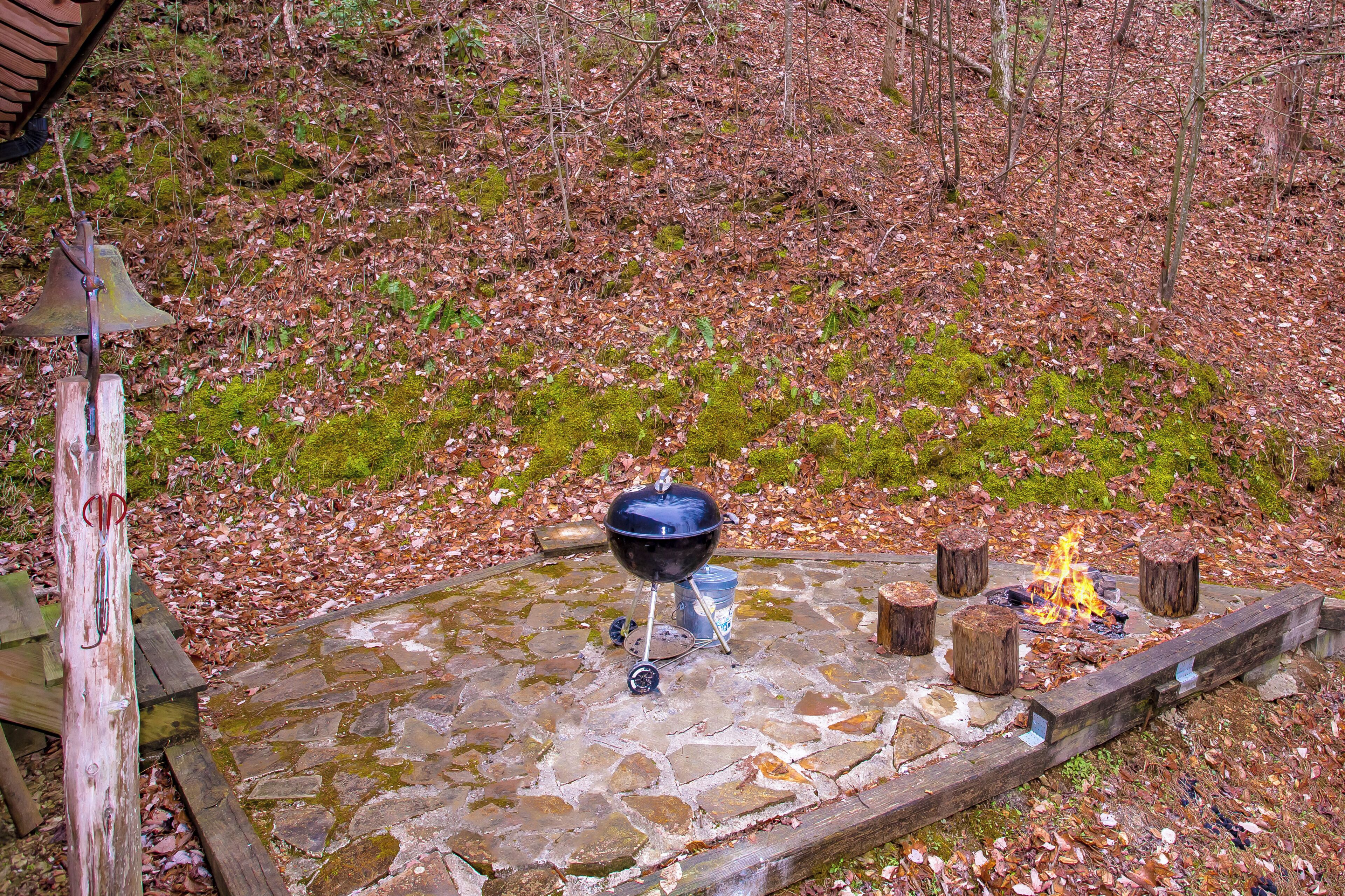 A stone fire pit with a grill and logs, surrounded by a forested area.