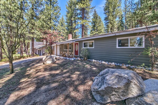 A single-story house with a dark siding exterior, a red door, and a small front yard with a large rock and a few trees.