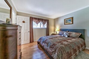 A bedroom with a bed, a dresser, a window with curtains, a nightstand with a lamp, and a framed picture on the wall.
