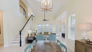 A spacious living room with a dining table, blue chairs, a sofa set, a staircase, and a chandelier. - Sea Shell Cottage~ Charming 3 BR/ 2-1/2 BA cottage! (Gulf Shores)