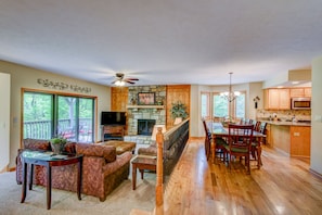 A spacious living room with a fireplace, a dining area with a large table, and a kitchen with wooden cabinets and a stone backsplash.