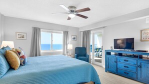 Une chambre à coucher avec un lit, un ventilateur de plafond, une télévision, une commode bleue et une vue sur l’océan.