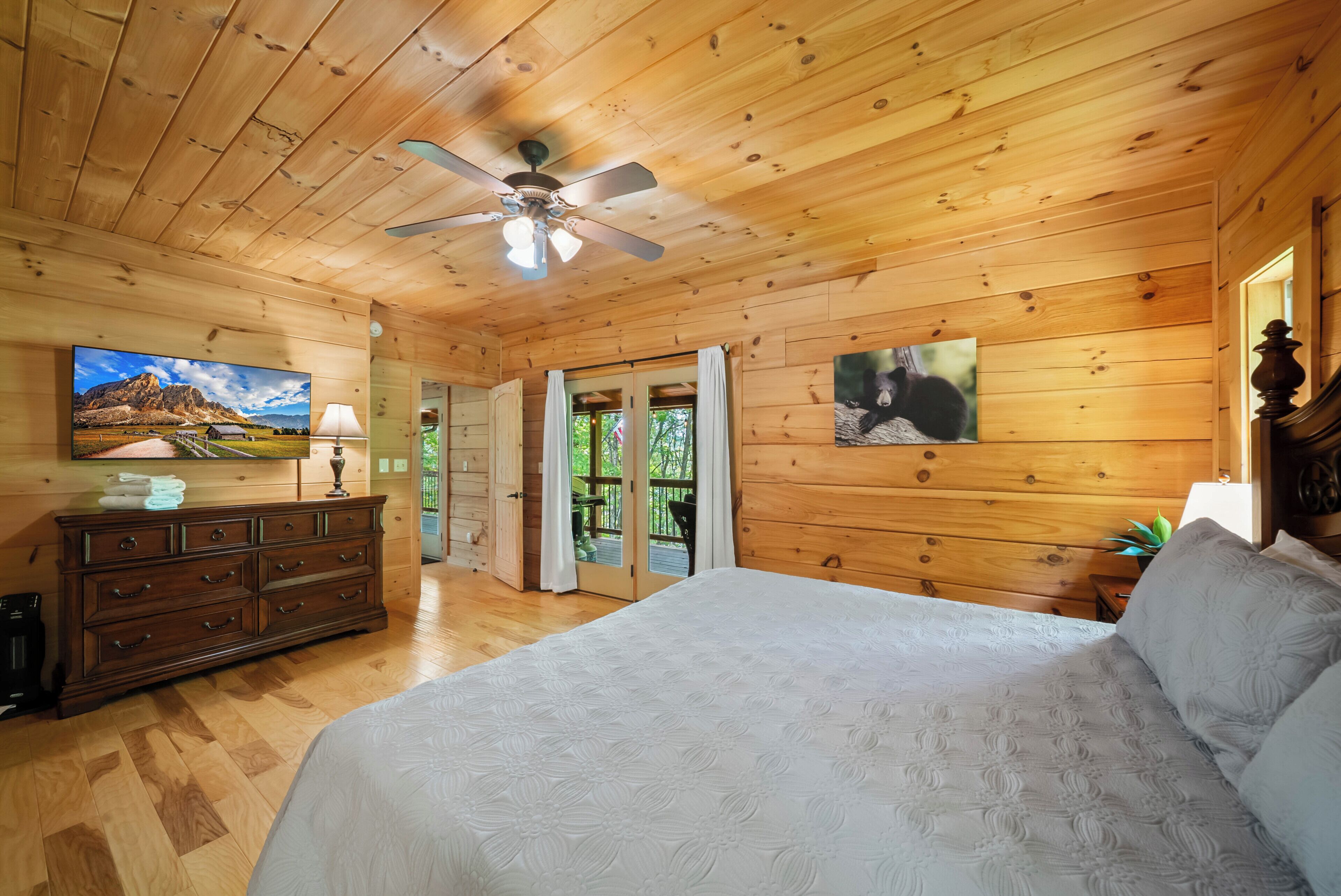A wooden cabin bedroom with a bed, dresser, ceiling fan, and two paintings.