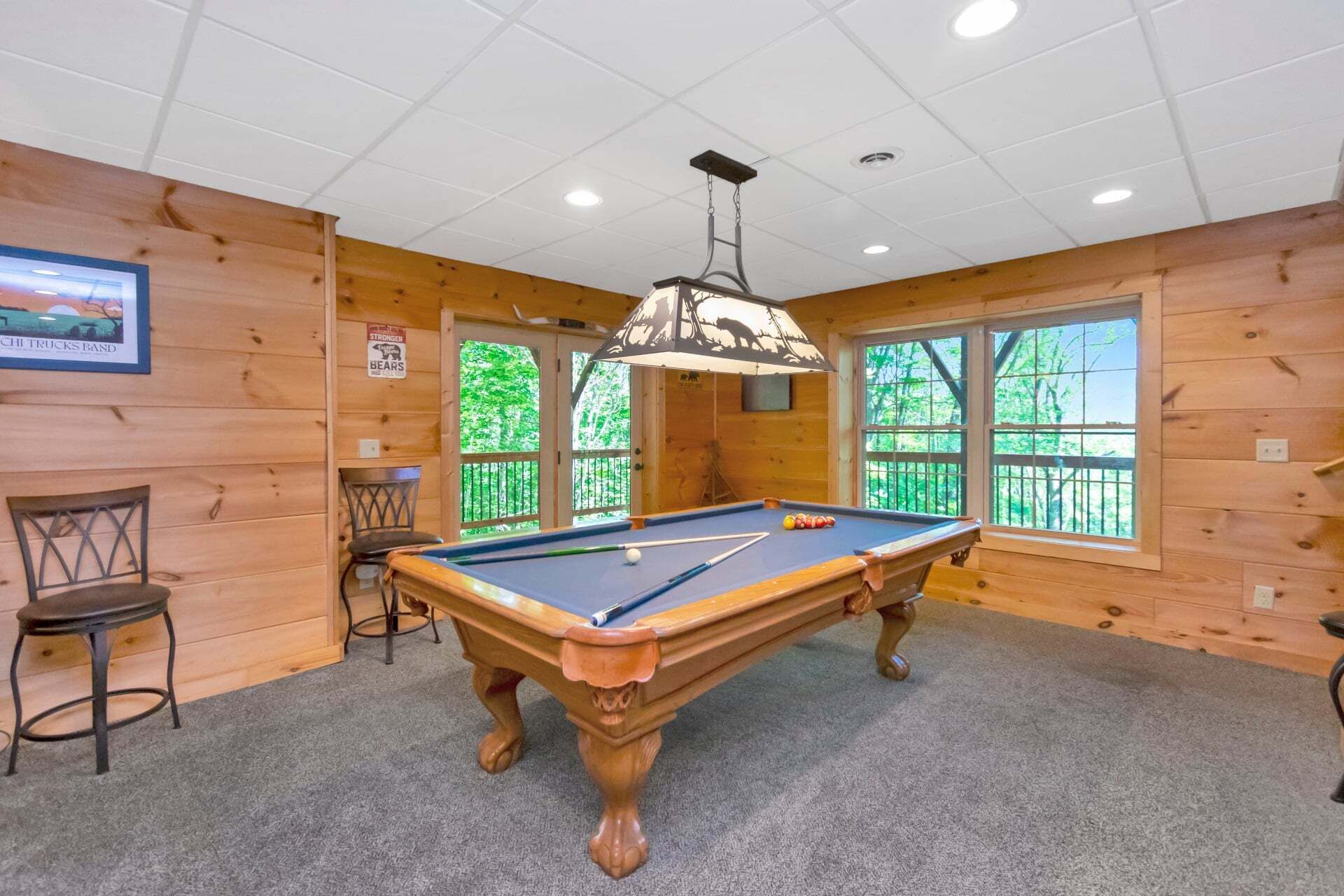 Une table de billard dans une pièce aux murs en bois, avec un ventilateur au plafond et des fenêtres donnant sur des arbres.