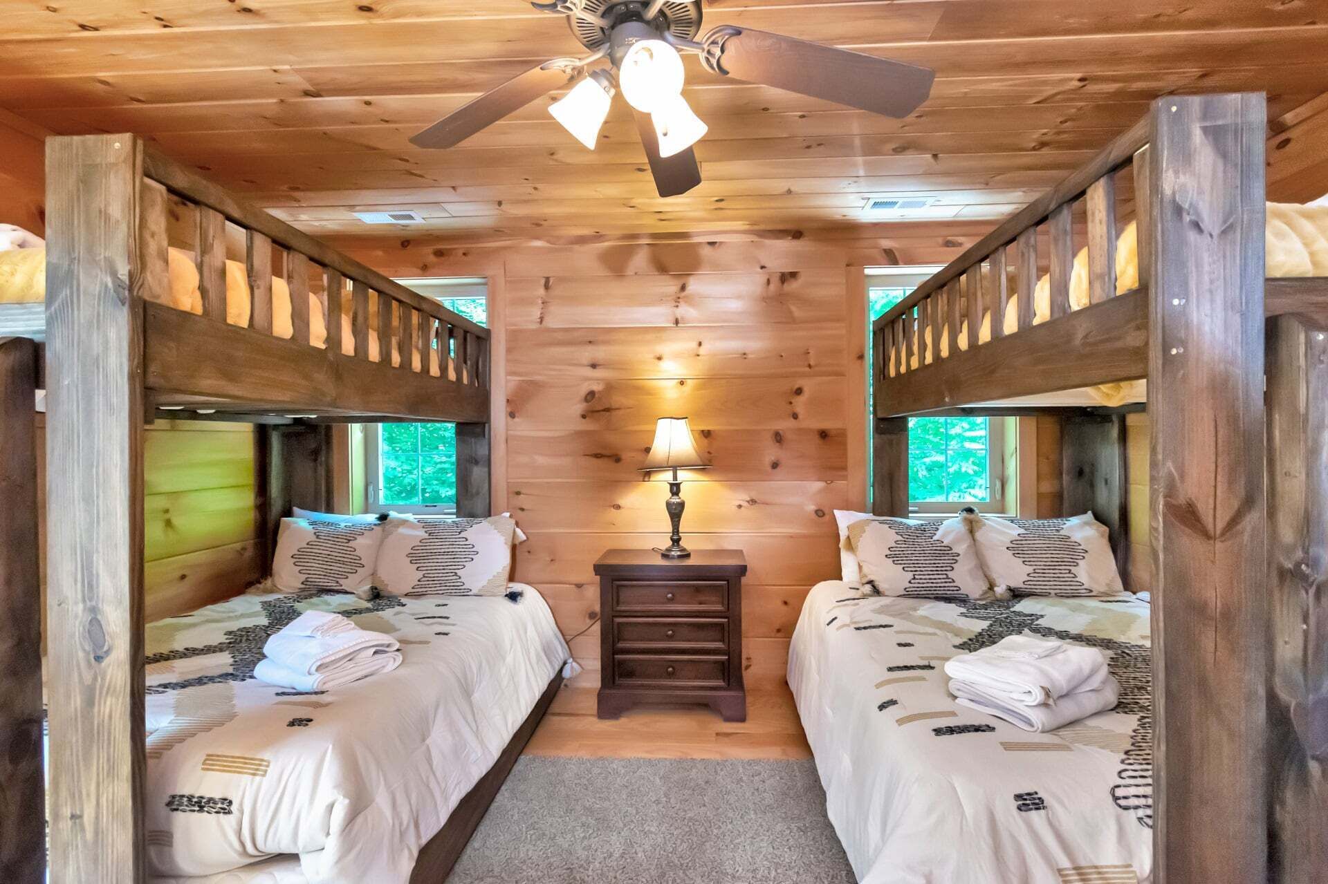A cabin-style room with two beds, a ceiling fan, a nightstand with a lamp, and a window.