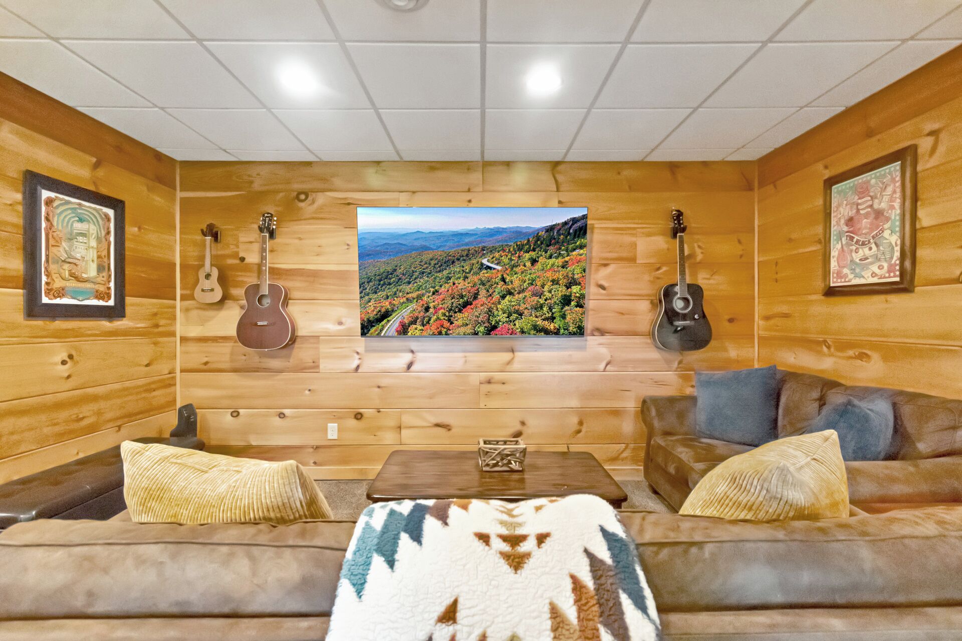 A cozy cabin interior with a wooden interior, a large TV screen, two guitars, a sofa, and a coffee table.