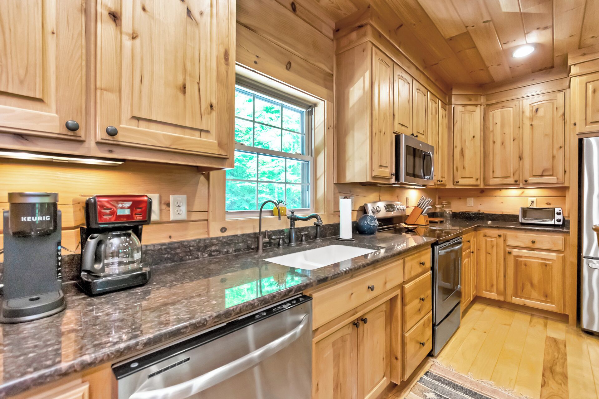 A kitchen with wooden cabinets, a granite countertop, a dishwasher, a microwave, a toaster, and a window with a view of greenery.