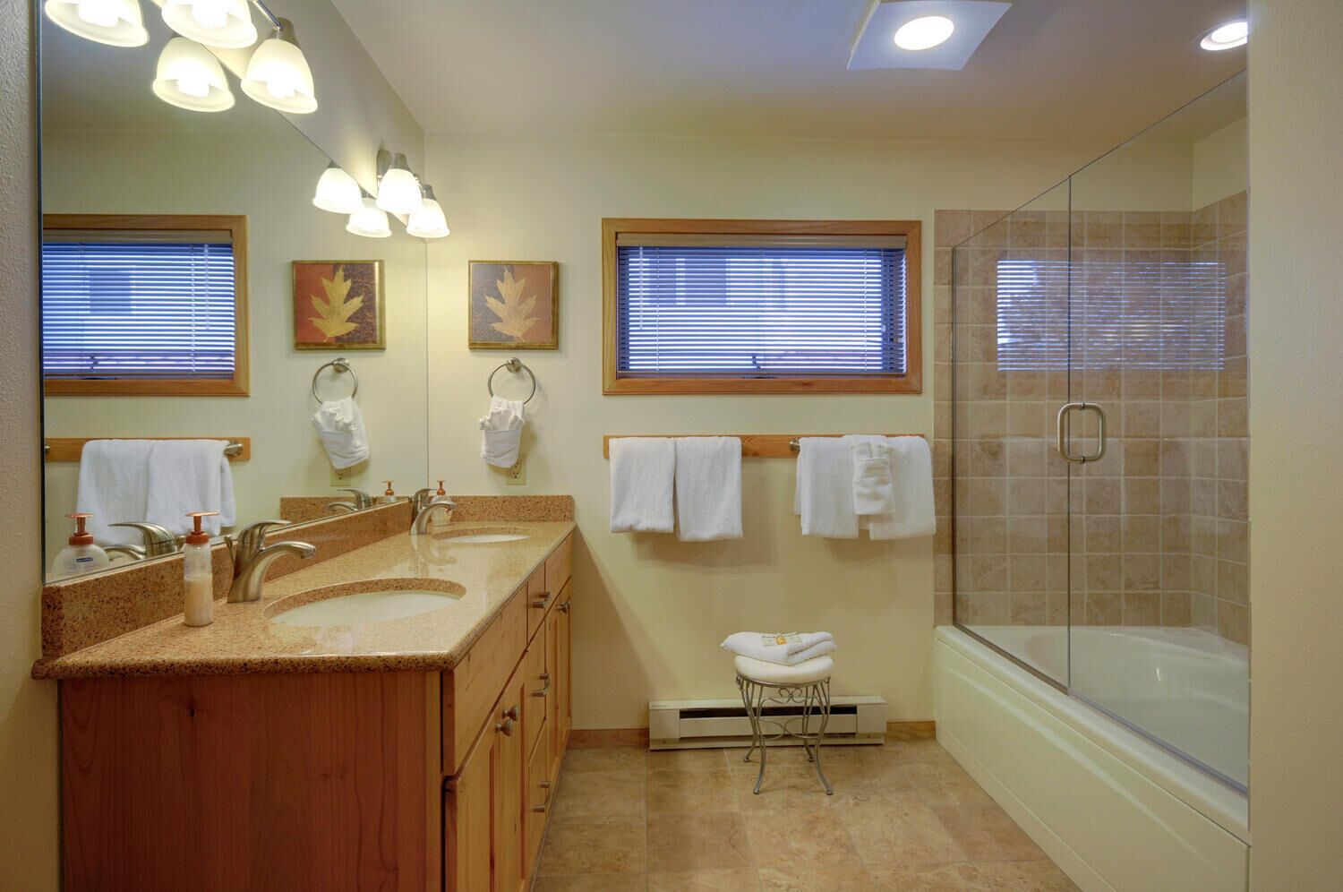 Une salle de bain avec un meuble-lavabo à deux vasques, une douche en verre et deux fenêtres équipées de stores.