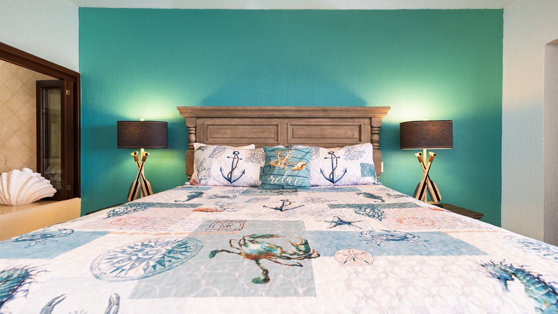 A bedroom with a teal wall, a wooden headboard, and a bed with a nautical-themed bedspread. There are two bedside tables with lamps and decorative items.