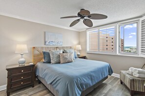 A bedroom with a large bed, two bedside tables with lamps, a ceiling fan, a window with blinds, and a view of the ocean. - Beachfront Views + Amazing Amenities – Islander 512 (Destin)