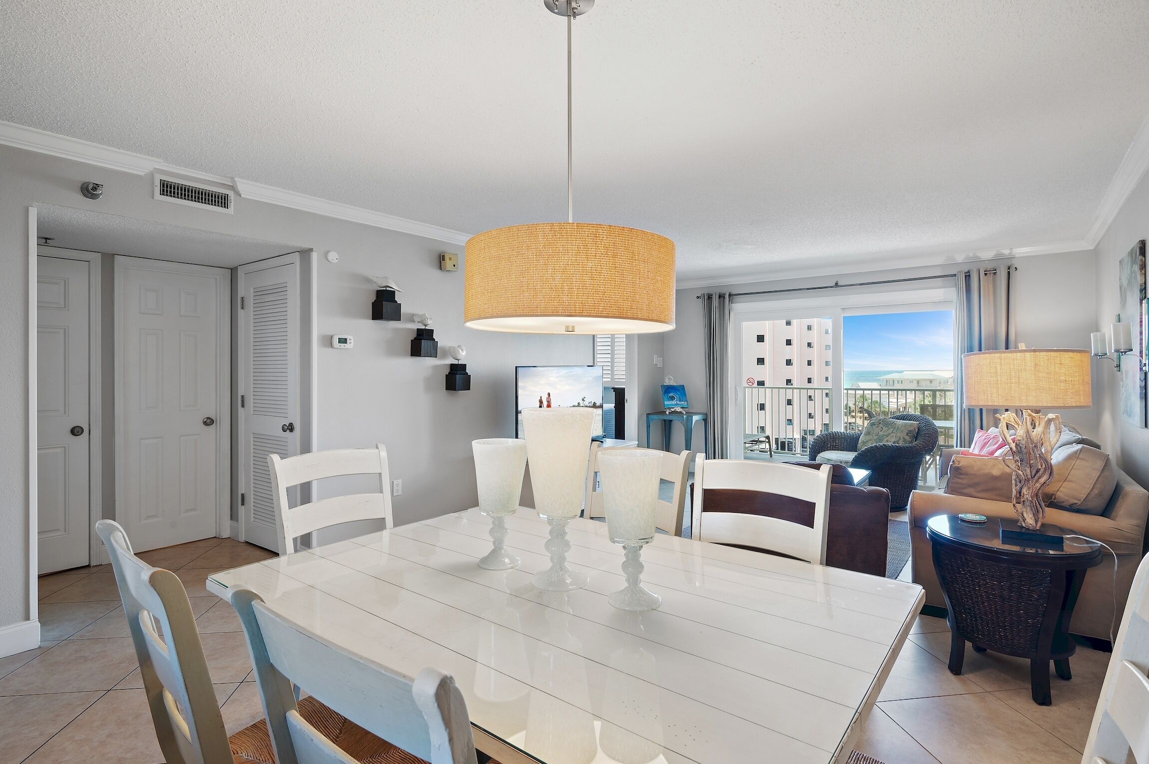 A modern living room with a dining area, featuring a large white table, white chairs, a wicker sofa, and a view of buildings through the window.