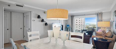 A modern living room with a dining area, featuring a large white table, white chairs, a wicker sofa, and a view of buildings through the window.