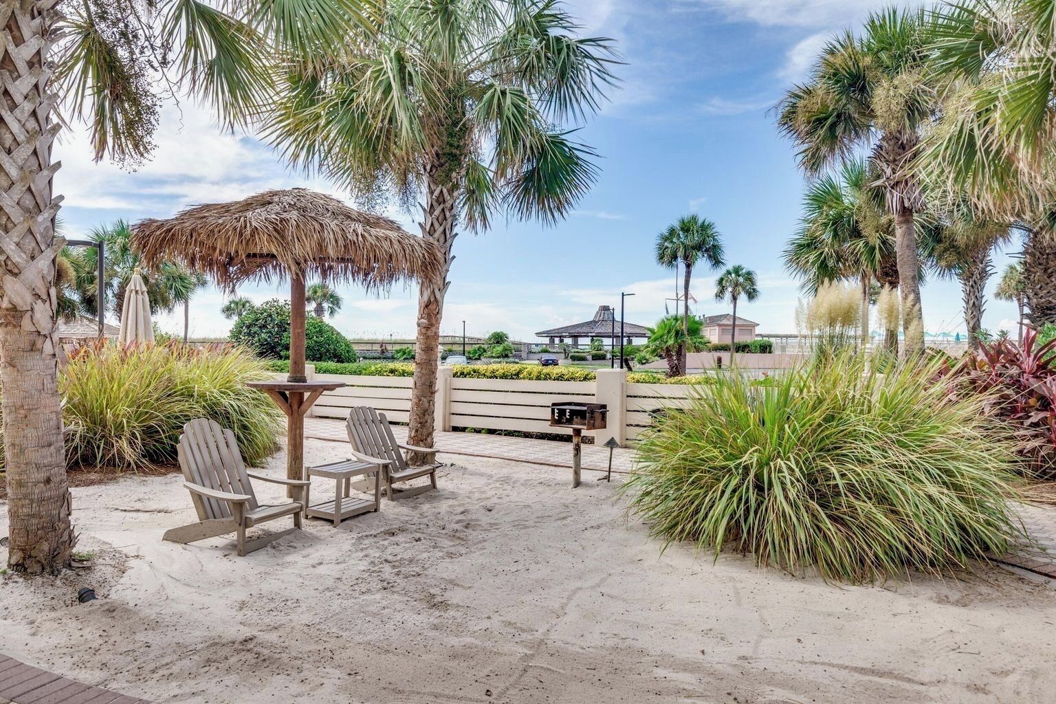 A beachside patio with wooden chairs, a thatched umbrella, and palm trees.