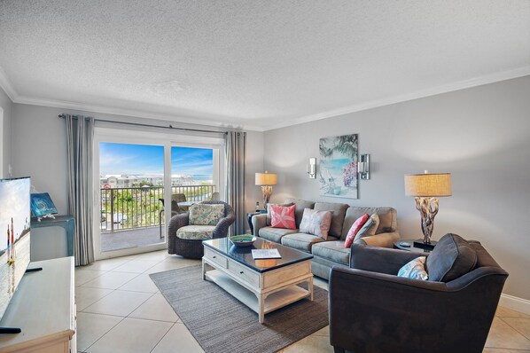 A modern living room with a sofa, coffee table, and a TV. There is a balcony with a view of the city. - Beachfront Views + Amazing Amenities – Islander 512 (Destin)