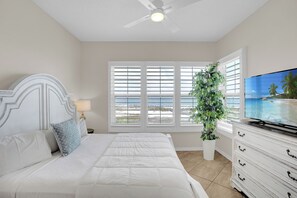 Une chambre à coucher comprenant un lit, un téléviseur, un ventilateur de plafond, une fenêtre avec des stores et une plante en pot.