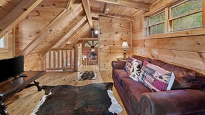 Interior de una cabaña de madera con techo de madera, un sofá de cuero, un televisor y un tapiz de piel de vaca.