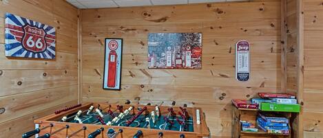 Una habitación con mesa de fútbolín, pared decorada con ilustraciones de la Ruta 66 y gasolineras, y estante con juegos de mesa.