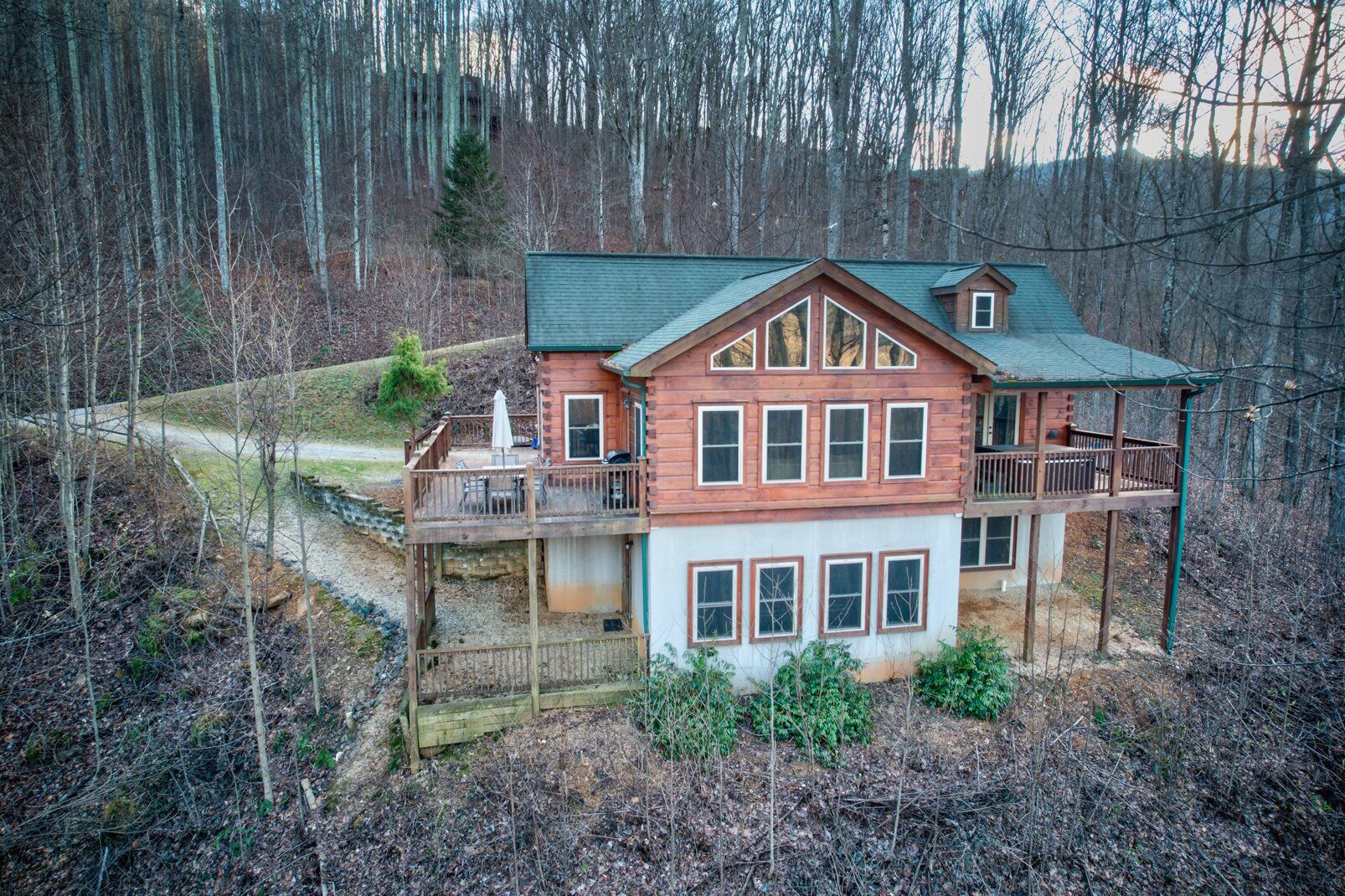 A two-story log cabin with a balcony and a deck, surrounded by a forested area.