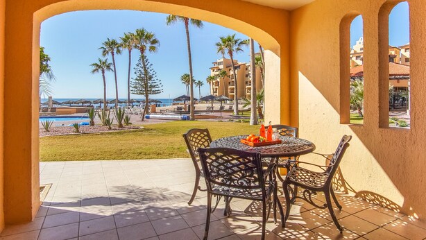 Una terrazza con tavolo e sedie, affacciata su una piscina e palme, con edifici sullo sfondo.