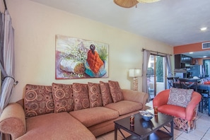 A living room with a sofa, a coffee table, and a chair. There is a painting on the wall and a kitchen area visible through an open door.