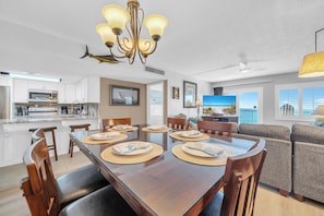 A modern kitchen with white cabinets, a dining area with a wooden table set for six, and a living room with a gray sofa and a television. - 720 -Breathtaking Sunset Views, Walk-in Shower, FREE beach service (Destin)