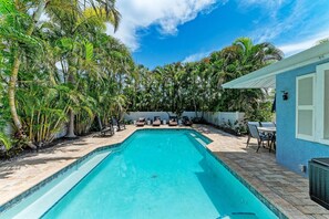 Una piscina rectangular con borde de azulejos, rodeada por un patio amoblado con muebles de exterior y una mesa.