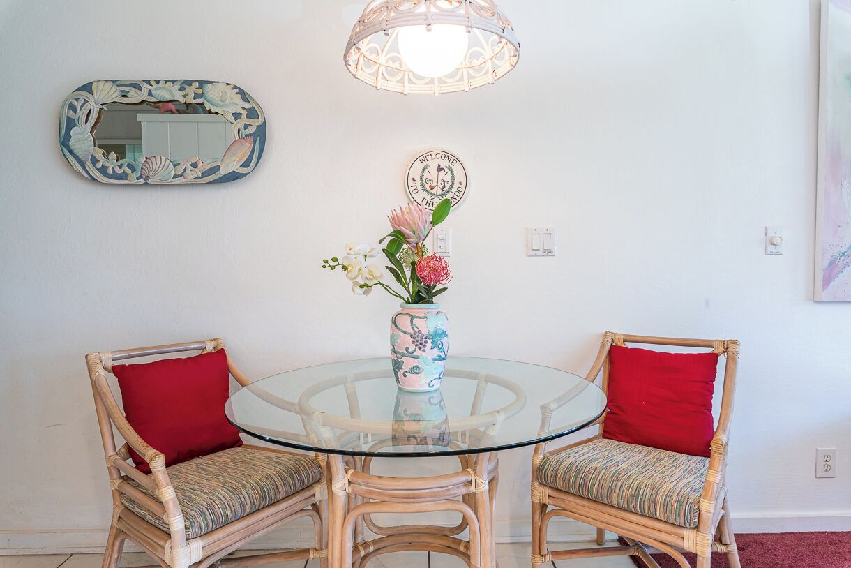 Una mesa redonda de vidrio con dos sillas de mimbre, cada una con un cojín rojo. Un jarrón con flores está sobre la mesa.