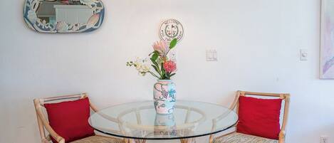 Una mesa redonda de vidrio con dos sillas de mimbre, cada una con un cojín rojo. Un jarrón con flores está sobre la mesa.