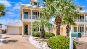 A two-story residential building with a covered front porch, a garage, and a well-maintained garden with palm trees and shrubs.