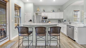 A modern kitchen with a central island, bar stools, white cabinets, stainless steel appliances, and a view through the window.
