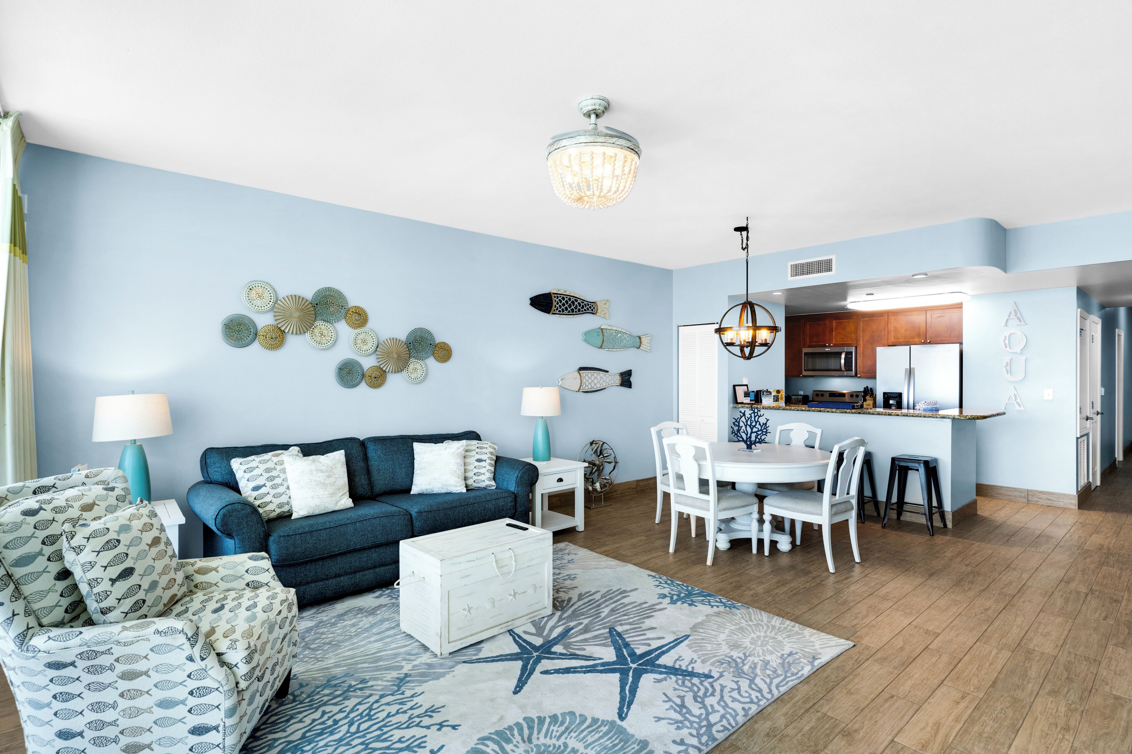 Una sala de estar con un sofá azul, una mesa de centro y un tapete con estampado de estrella de mar. Hay una zona de comedor con una mesa y sillas blancas, y una cocina con armarios de madera y un refrigerador blanco.