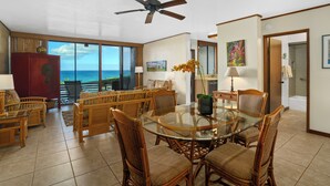 A spacious living area with a glass-top dining table surrounded by wicker chairs, a ceiling fan, and a view of the ocean. - Poipu Makai B-1, A/C - Oceanfront - Enjoy the view! (Poipu)