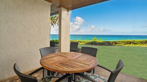 A covered patio with a round wooden table and wicker chairs, overlooking a grassy area and the ocean. - Poipu Makai B-1, A/C - Oceanfront - Enjoy the view! (Poipu)