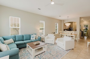 A spacious living room with a blue sofa, white armchairs, a wooden coffee table, and a dining area with white chairs and a table.