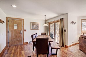 A dining area with a wooden table and chairs, a ceiling light, and a view through a glass door to an outdoor stone wall.