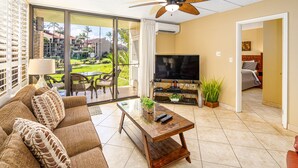 Una sala de estar con sofá, mesa de centro, televisor y un balcón con vista a un jardín y edificios.