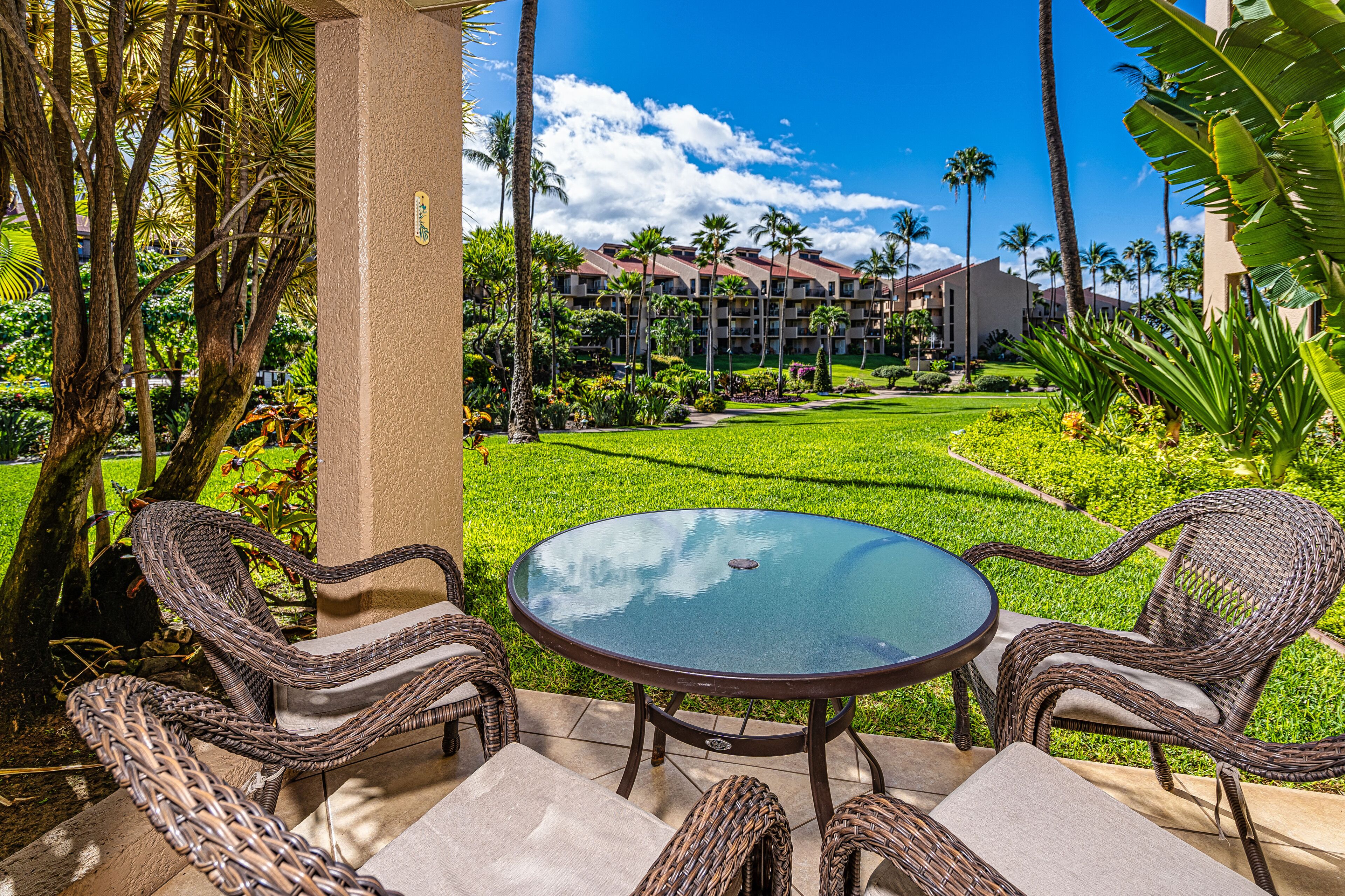 An outdoor seating area with wicker chairs and a glass table, overlooking a well-maintained lawn and a building with multiple balconies.