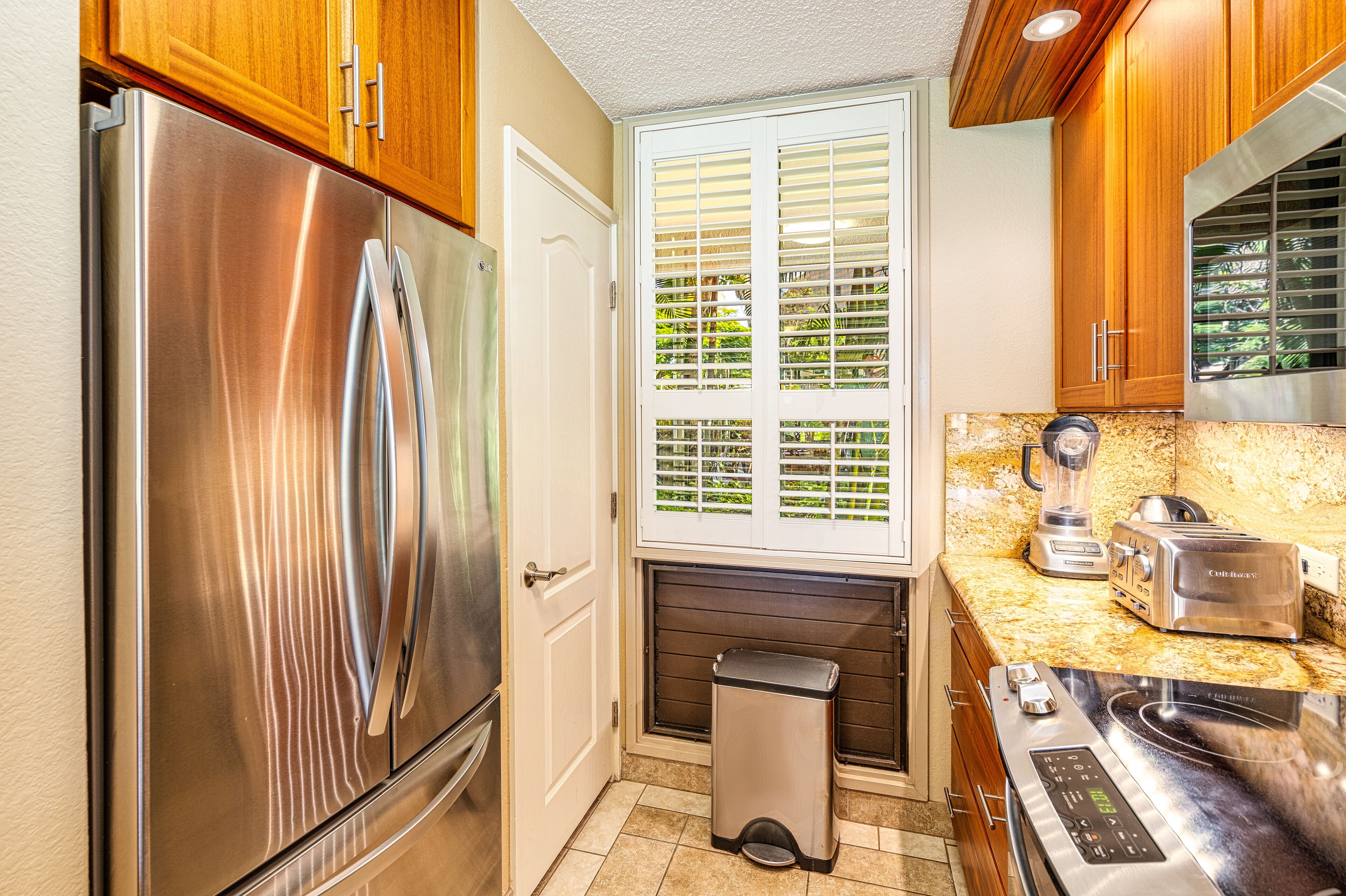 Una cocina compacta con refrigerador de acero inoxidable, horno empotrado, encimera con licuadora y tostadora, y ventana con persianas.