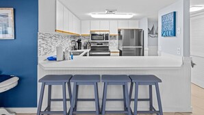 A modern kitchen with white cabinets, a central island with bar stools, a refrigerator, and a microwave. - Unit 102 - Beautiful beachfront 2-BR - Newly Remodeled w/ Washer&Dryer (Destin)
