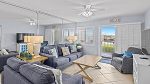 A spacious living room with a grey sofa set, a wooden coffee table, and a TV. There is a ceiling fan, a large window with shutters, and a view of the outdoors. - Unit 102 - Beautiful beachfront 2-BR - Newly Remodeled w/ Washer&Dryer (Destin)