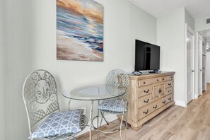 Ein Zimmer mit Fernseher auf einer Holzkommode, einem Glastisch mit zwei Stühlen und einem Bild an der Wand.