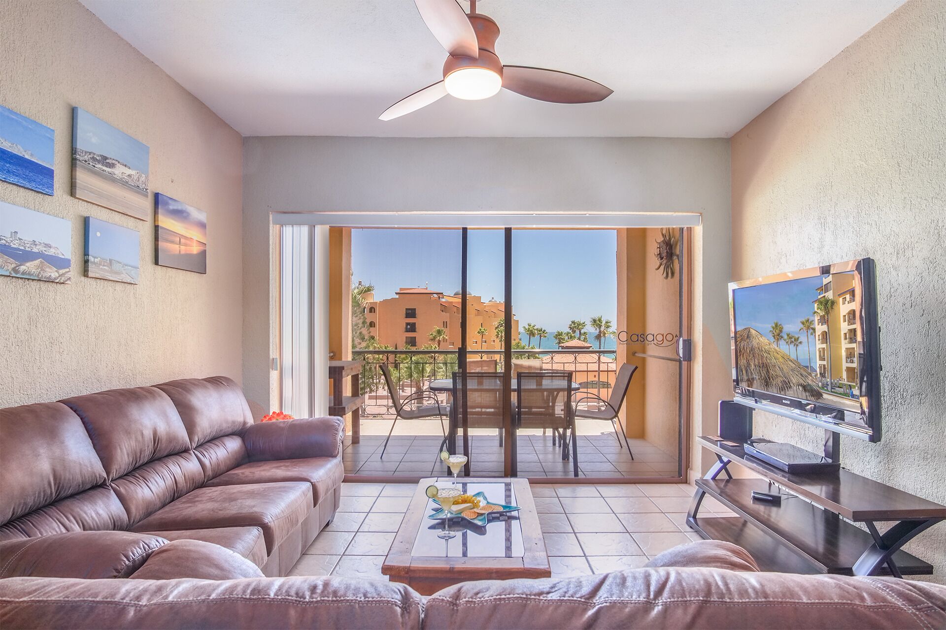 A living room with a brown leather sofa, a coffee table with a drink, a TV on a stand, and a balcony with a view of buildings and palm trees.