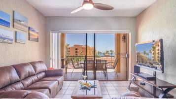 A living room with a brown leather sofa, a coffee table with a drink, a TV on a stand, and a balcony with a view of buildings and palm trees.