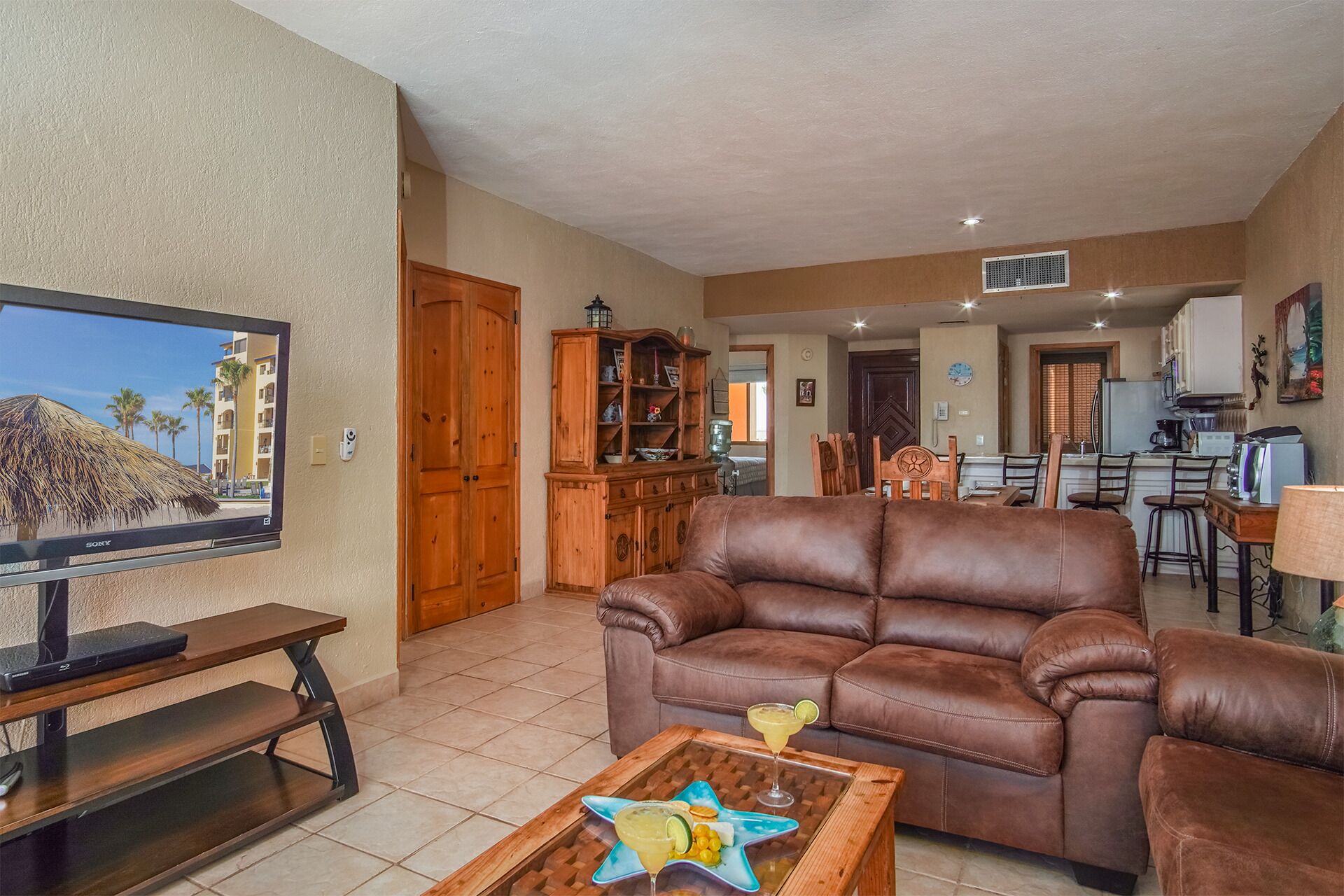A living room with a brown leather sofa, a coffee table with drinks, a TV on a stand, wooden cabinets, and a kitchen area in the background.