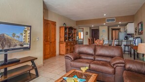Ein Wohnzimmer mit braunem Ledersofa, einem Couchtisch mit Getränken, einem Fernseher auf einem Standfuß, Holzschränken und einer Küche im Hintergrund.