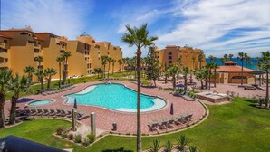 Ein Resort mit einem zentralen Schwimmbecken, umgeben von Palmen und Liegestühlen, im Hintergrund mehrere Hotelgebäude.