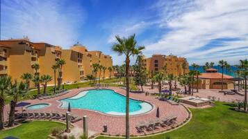 A resort with a central swimming pool surrounded by palm trees and lounge chairs, with multiple hotel buildings in the background.