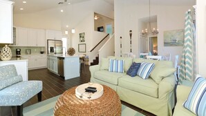 A spacious living room with a light green sofa, a wicker coffee table, and a blue patterned armchair. A kitchen with white cabinets and a stainless steel refrigerator is visible in the background.