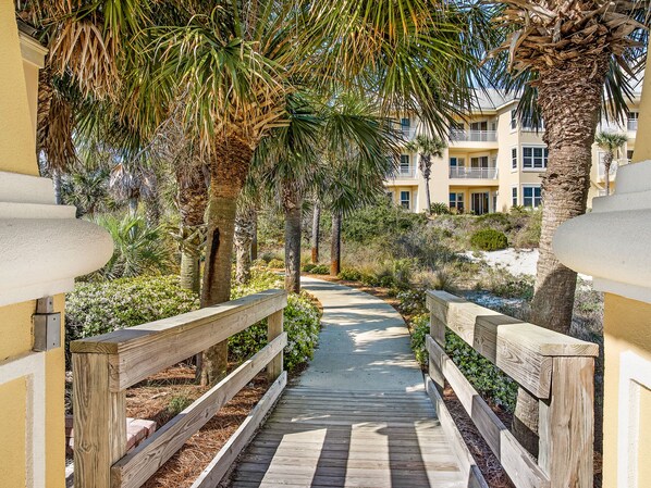 A wooden pathway leading to a building with balconies, surrounded by palm trees and greenery.